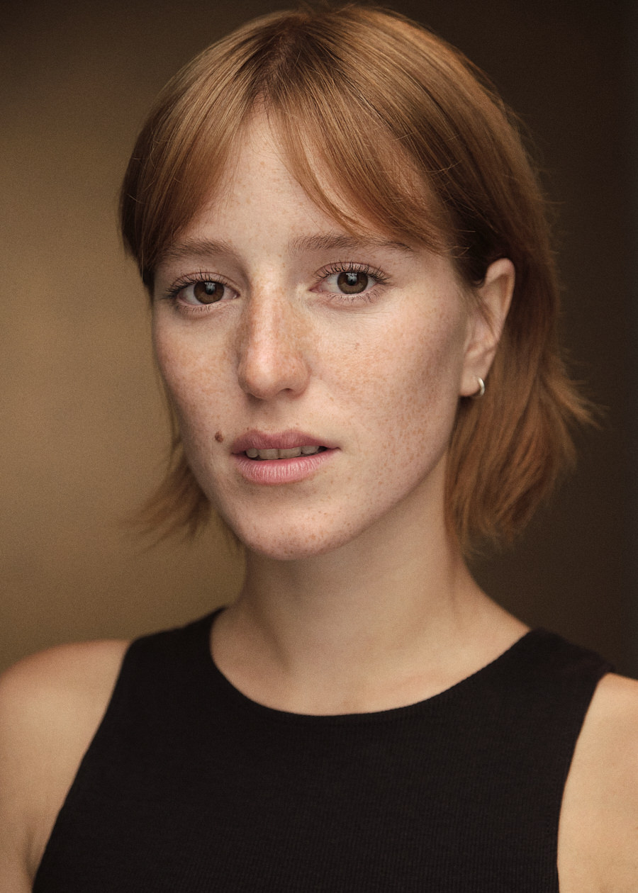Headshot of German actress Leah Lavinia, distinguished by her striking red hair, freckles, and light brown eyes. Her intense gaze is fixed upon the camera as she wears a black vest, exuding confidence and charisma.