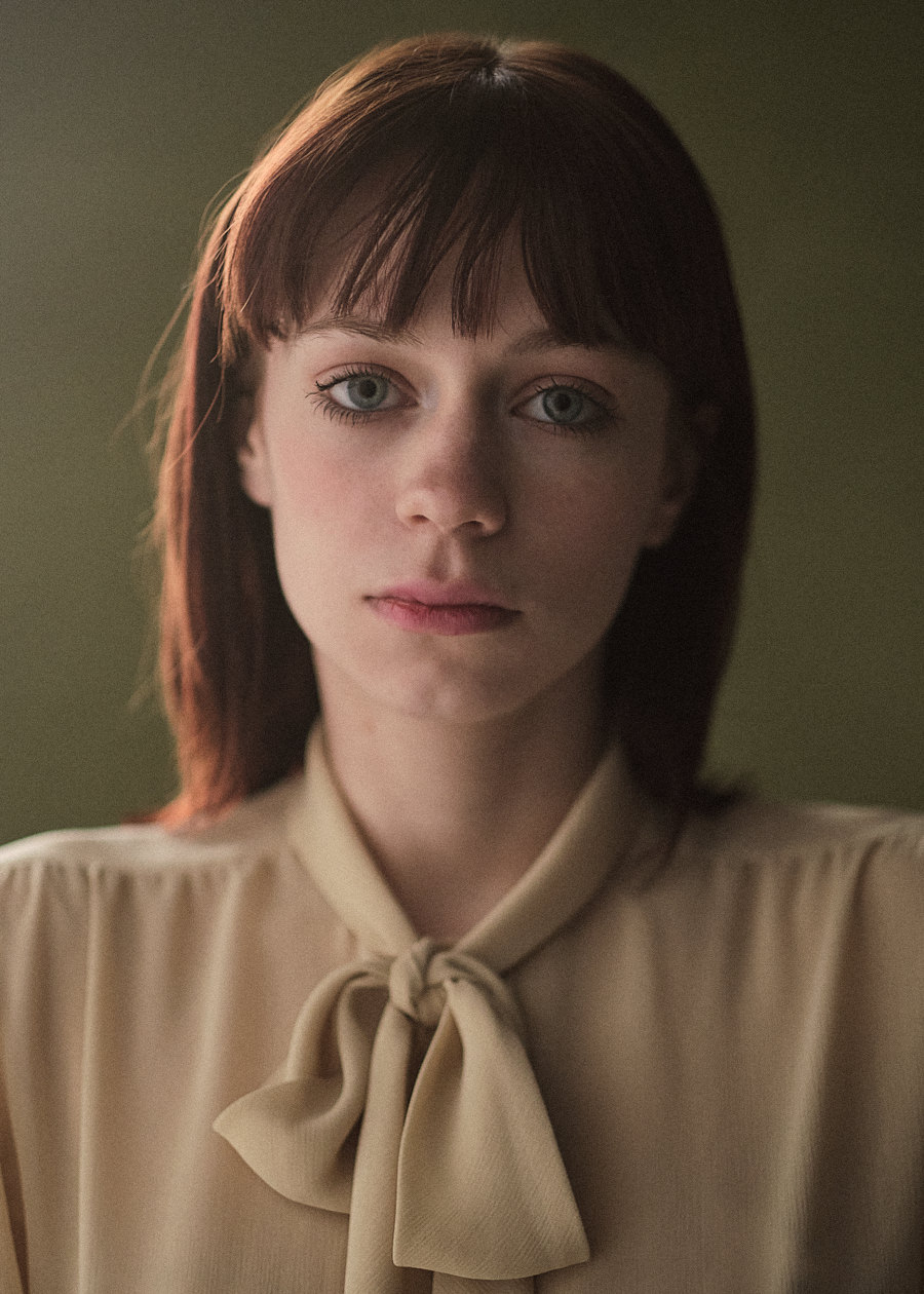A compelling actors headshot of a young woman with vibrant red hair and strikingly expressive large blue eyes, emotionally gazing directly into the camera. She's dressed in a beige shirt, set against an olive green background.