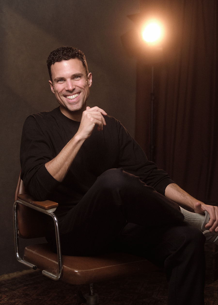 Professional portrait of Alejandro Lopez, sitting on a 70s office chair and smiling naturally. There is a studio light behind him creating a lens flare in the camera.