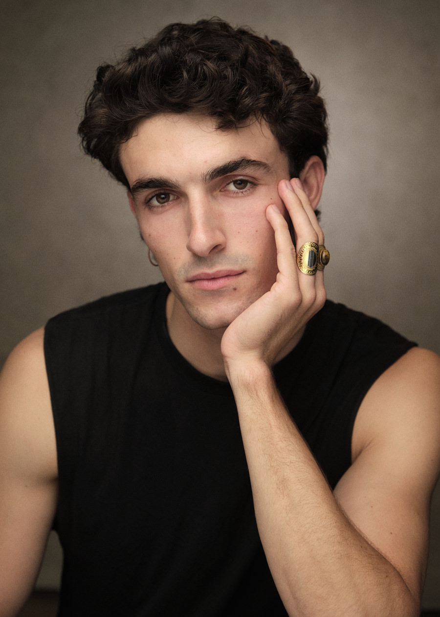 Actors headshot of Spanish actor and model Alex Peral, showcasing his striking black wavy hair, dark eyes, and strong features. Seated with his hand on his chin, he's dressed in a sleeveless black T-shirt and adorned with stylish gold rings on his fingers.