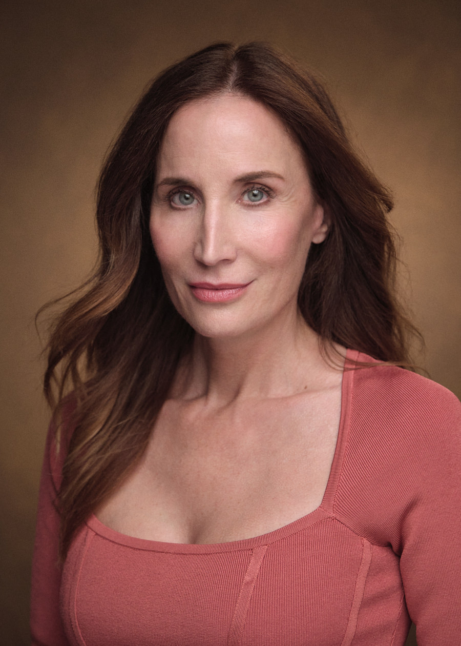Professional headshot of American actress Anne Alexander Sieder, featuring long, wavy brown hair and captivating blue eyes. She exudes confidence with a bright smile while wearing a stylish red top. The backdrop provides a textured, warm brown setting.