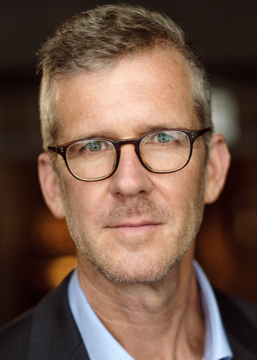A corporate portrait photo of Benjamin Newland, founder of Newland Ventures LLC. He's has short light hair and is wearing glasses. He's looking directly into the camera confidently and has a confident half smile.