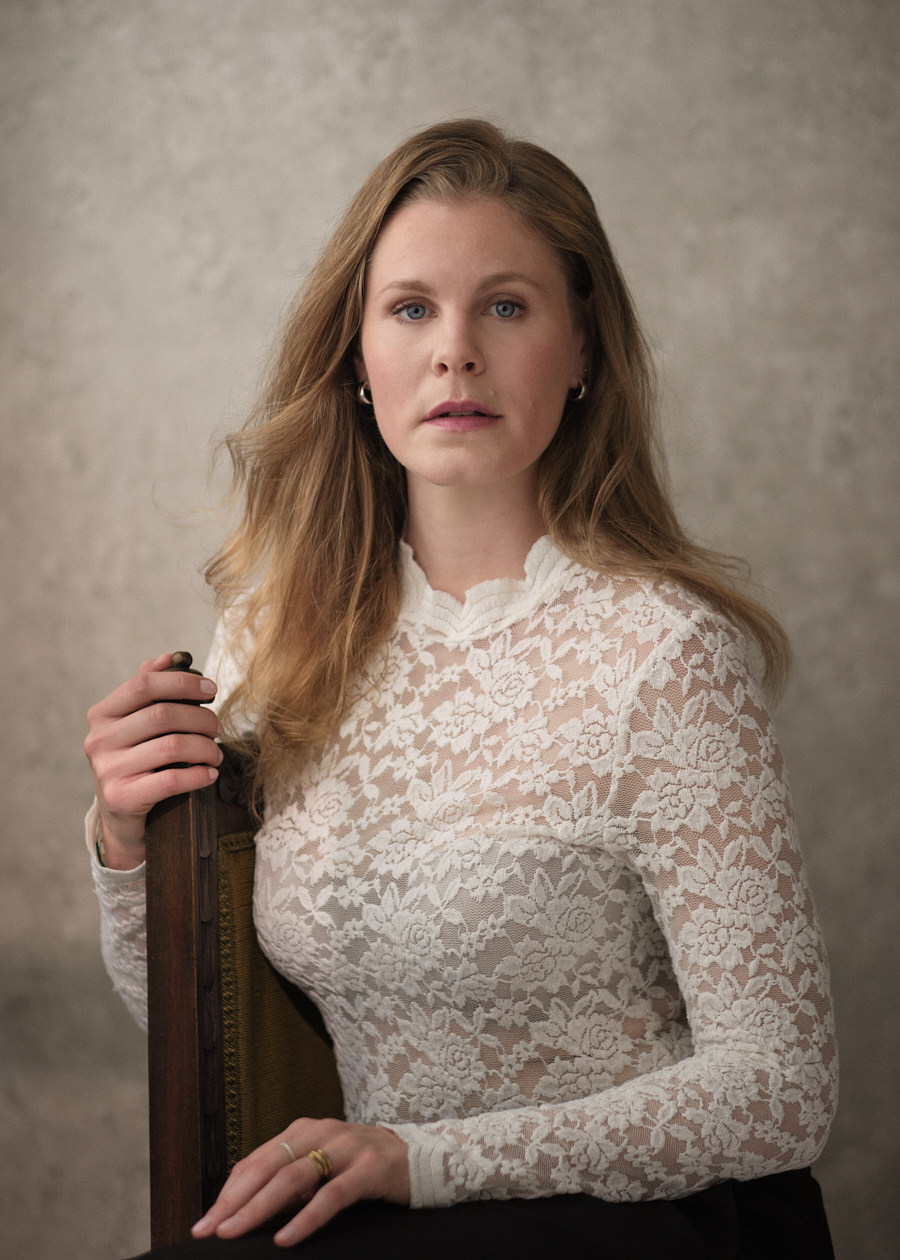 Professional portrait of German actress Charlotte Mednansky, a youthful talent with wavy blonde hair. She gracefully poses on an antique chair, adorned in a cream lace top, gazing gently into the camera. The textured cream background evokes the ambiance of a classic Renaissance painting.