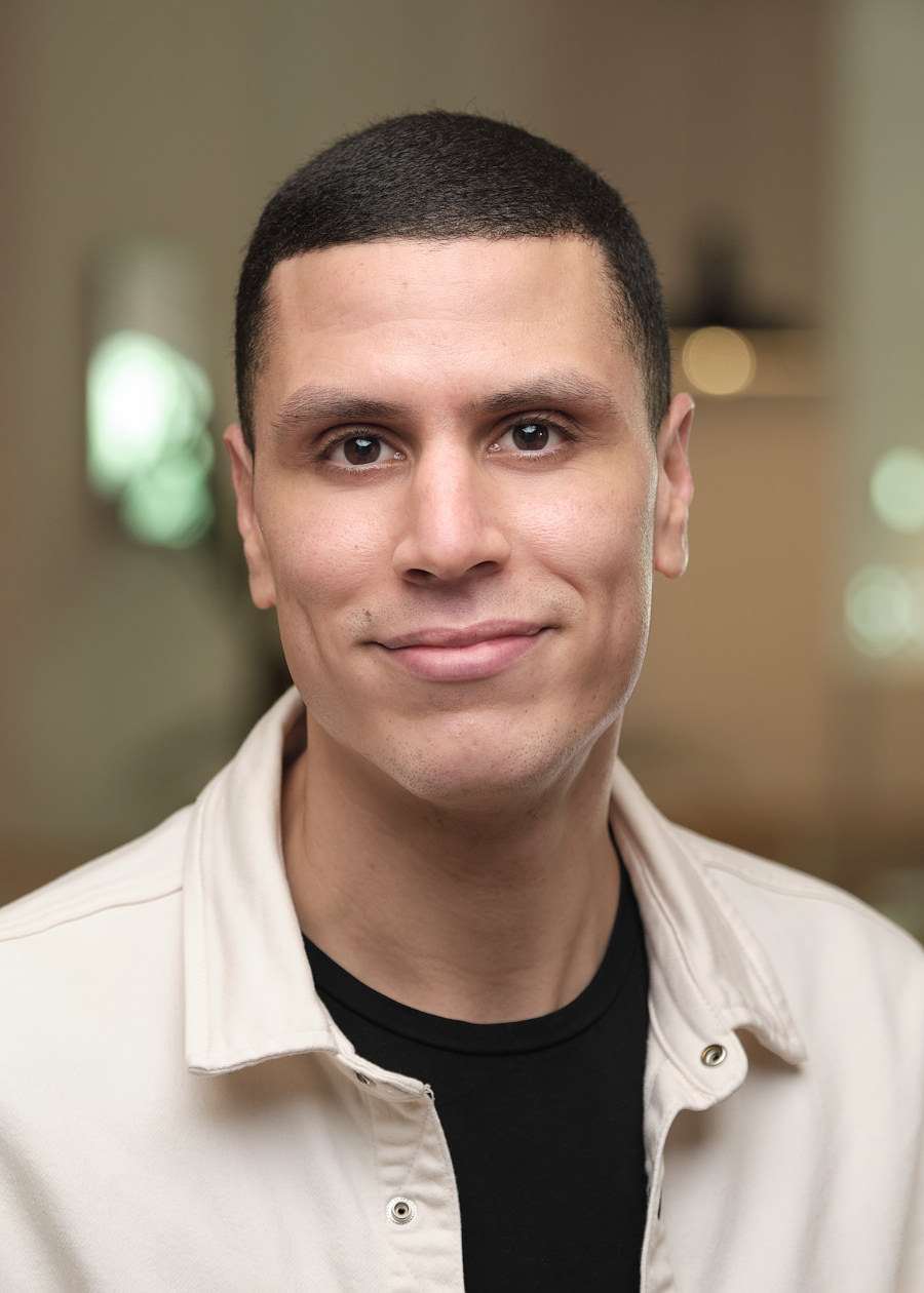 A professional headshot of a team member from Mixpanel. He is French and has short dark hair and dark eyes. He's smiling directly at the camera. He's wearing a beige shirt with a black Tshirt underneath and looks very fashionable. The background of the photo is a blurred office with lights shining.