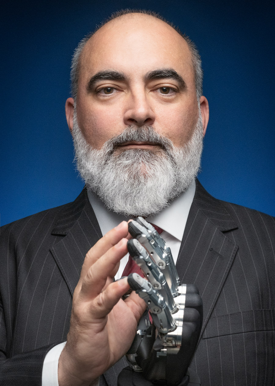 An exceptional professional portrait of Javier Garcia, the CEO of SCHUNK. This striking image captures a man with a robotic hand, sporting a bald head and a grey beard, elegantly dressed in a formal suit. Set against a deep blue background, he raises one hand in a gesture of authority, while his other hand is a testament to innovation with its robotic enhancement.