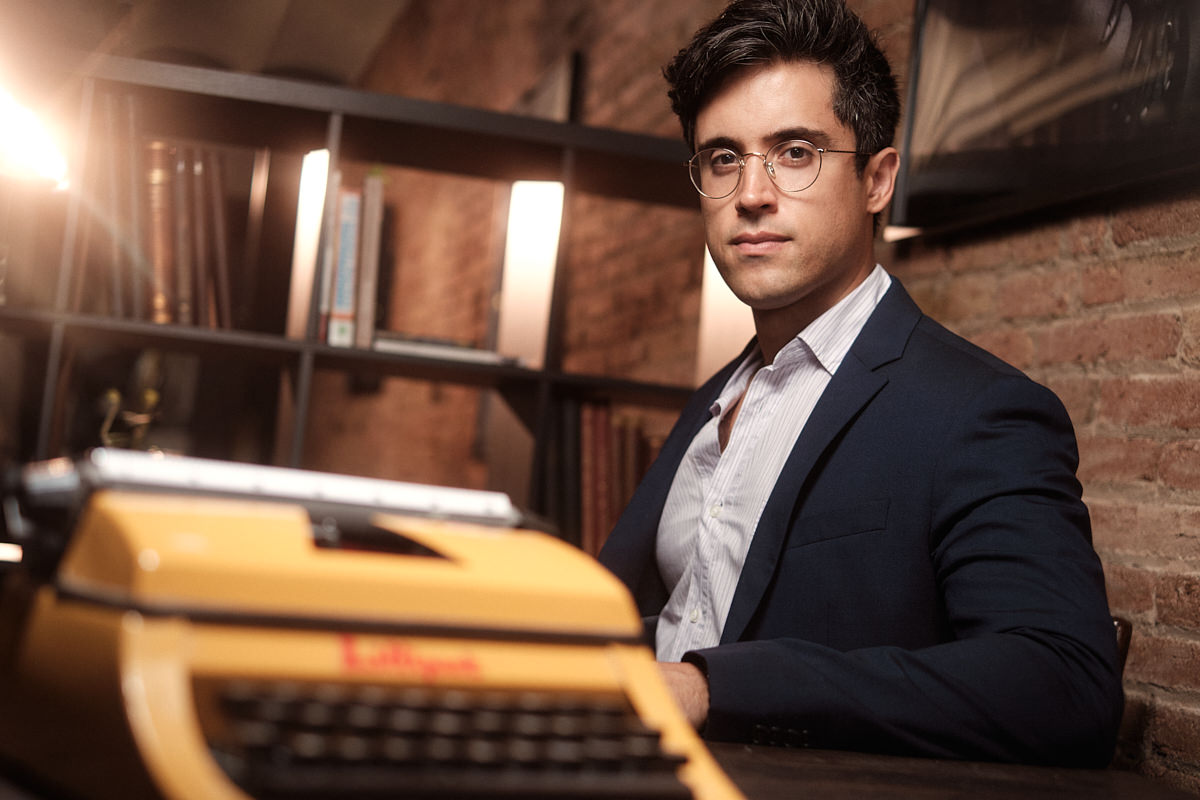 Modern corporate photography of Jose Camero. He's a young author, sitting at a table with a book shelf in the background, and an out of focus vintage typewriter in the foreground. He's wearing a suit and glasses.