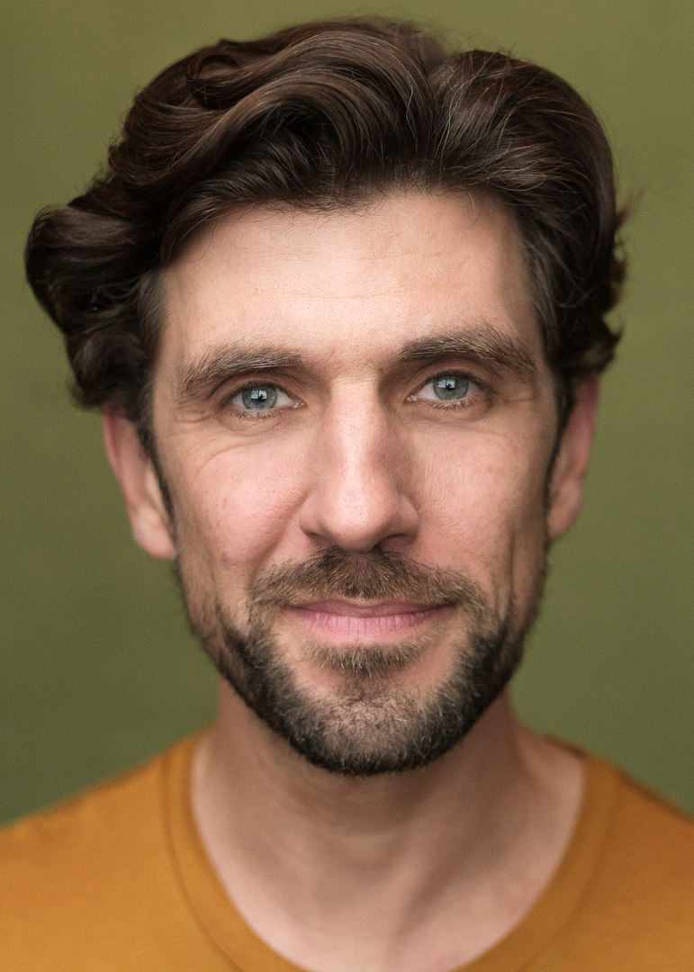 A captivating headshot of Belgian actor Julien Paschal, showcasing his confident gaze and expressive blue eyes as he looks directly into the camera. He's attired in an orange T-shirt against a vibrant green background.