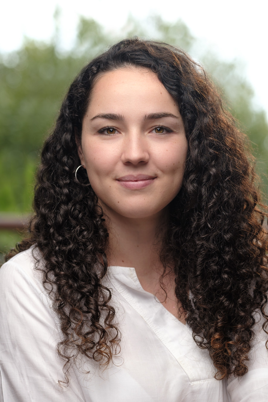 A corporate headshot of a young Spanish woman with curly brown hair and light brown eyes. She's wearing a white shirt and smiling naturally. The background of the photo is blurred and of trees. The lady works for Lemonade Software Development in Barcelona.