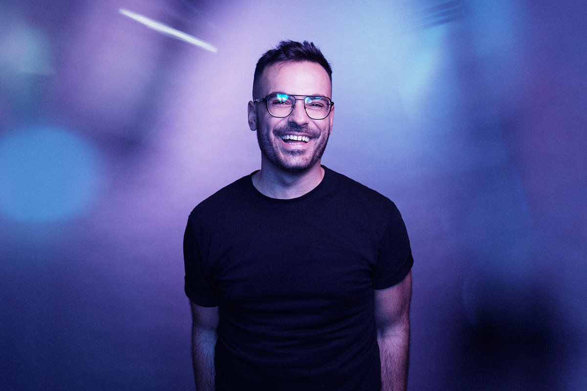 A captivating LinkedIn profile photo featuring a young designer with short brown hair and glasses. He exudes a welcoming demeanor, captured in a moment of genuine laughter. The image is infused with vibrant blue and purple lights, evoking a futuristic and modern vibe, setting a visually engaging tone for his professional online presence.