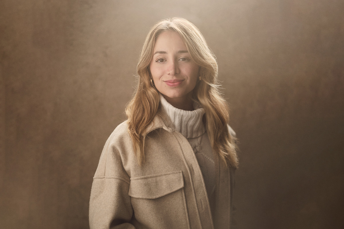 A polished LinkedIn photo showcasing a confident young South American woman. Her blonde hair beautifully complements her attire, consisting of a cream jumper paired with a stylish beige oversized jacket. Against a creamy backdrop, a gentle light flare from above adds a touch of radiance to the scene as she smiles with assurance, making an impactful professional statement.