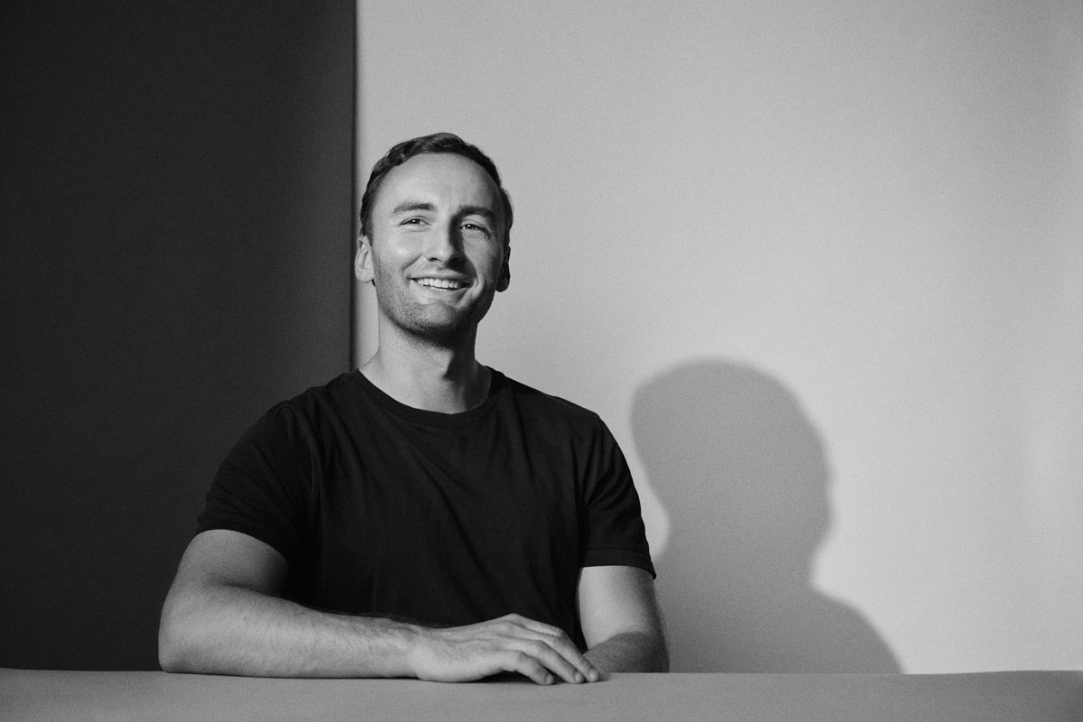 A Black and White professional portrait of a young designer sitting at a white table. He's looking off camera and is laughing. He has short brown hair.