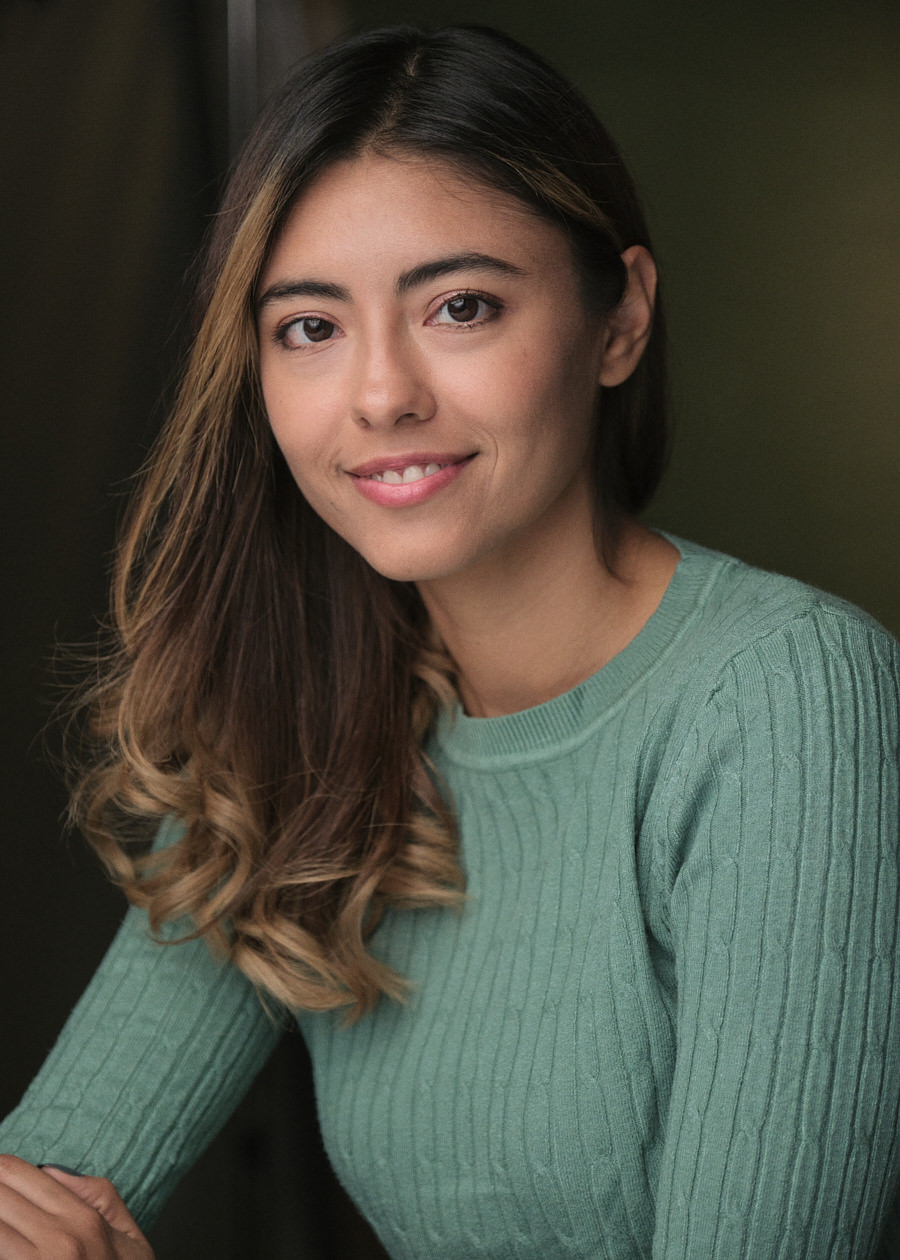 A professional headshot featuring the talented actress Mayela De L'arc, radiating warmth with a gentle smile directed at the camera. She's elegantly dressed in a green jumper, exuding charm and grace.
