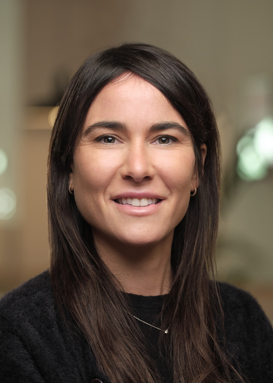 A professional headshot of a young Spanish lady. She works for the Barcelona team of Mixpanel. She has long dark hair and brown / green eyes. She's smiling very confidently. She's wearing a black jumper and the background of the photo looks like an out of focus office.