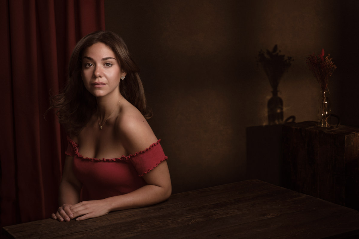A stylised portrait of Italian actress Misa D'Angelo, dressed in a striking red top, seated at an aged brown table. The backdrop features flowers and a red curtain, creating a moody ambiance in this captivating portrait.