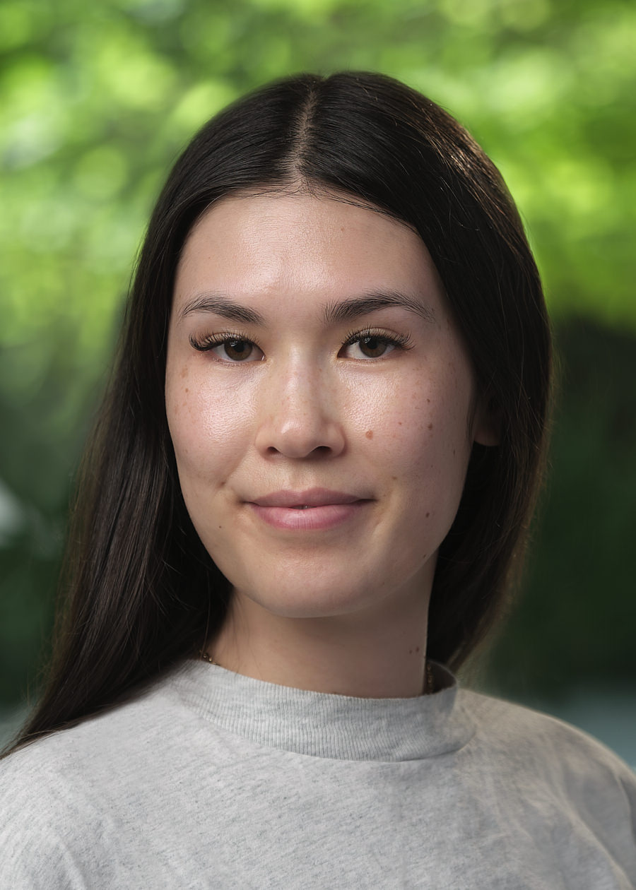 A professional headshot of a young Asian woman. She has long dark hair and dark eyes. She's wearing a grey jumper and is smiling. The background of the photo is very out of focus and green.