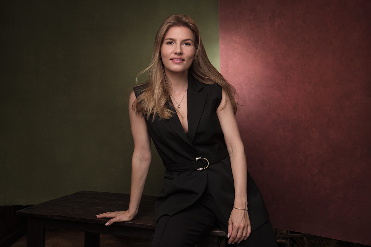 An example of a great LinkedIn photo. It has a young business woman sitting in a relaxed way on a vintage table. She's blonde and smiling in a relaxed way. She's wearing a modern black, sleeveless business suit.