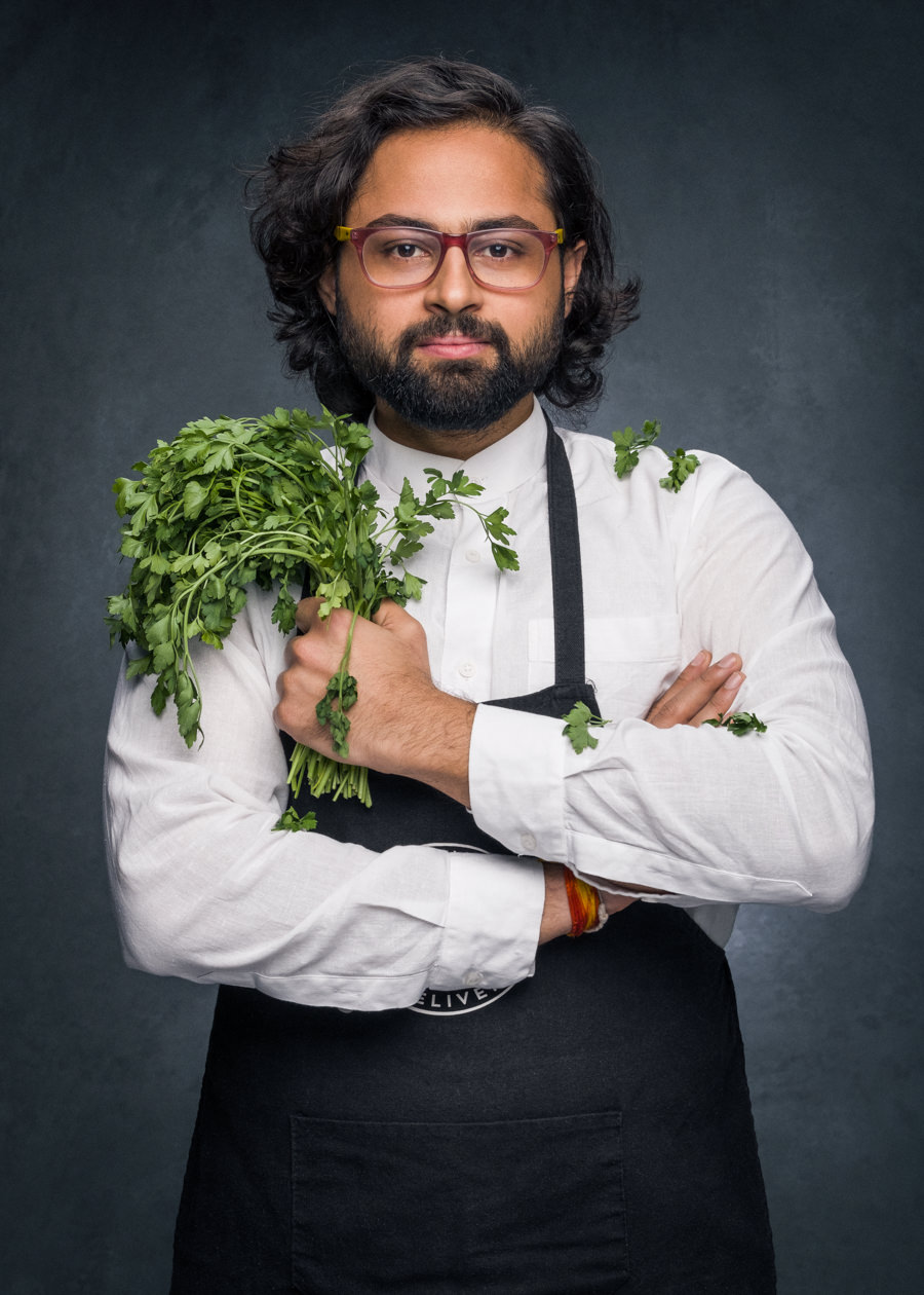 A professional portrait of an Indian chef, posing with a bunch of herbs in his hand. He has mid-length wave black hair, a black beard and glasses. He's wearing a chefs outfit with a black apron.