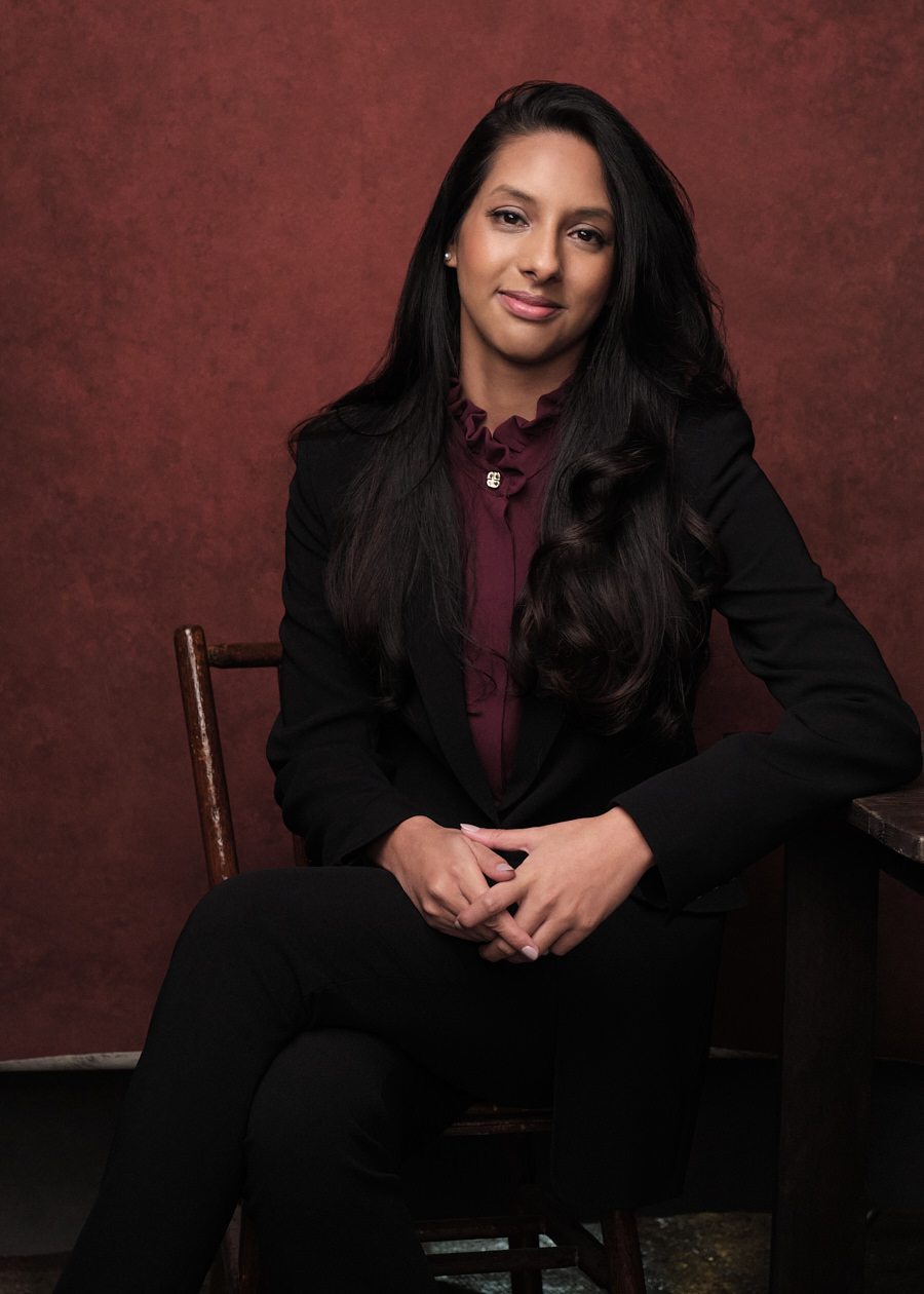 A professional portrait for Urmi Ahmed, a young Indian business woman, sitting on a wooden chair in a relaxed pose and smiling. She's wearing a modern black suit with a maroon shirt.