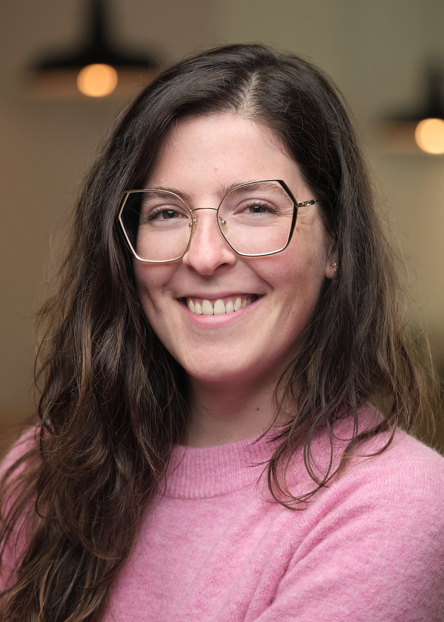 A professional headshot for a team member of Mixpanel. The headshot shows a young woman, with long dark wavy hair. She's wearing modern square glasses and a bright pink jumper. She looks very happy. The background shows an out of focus office.