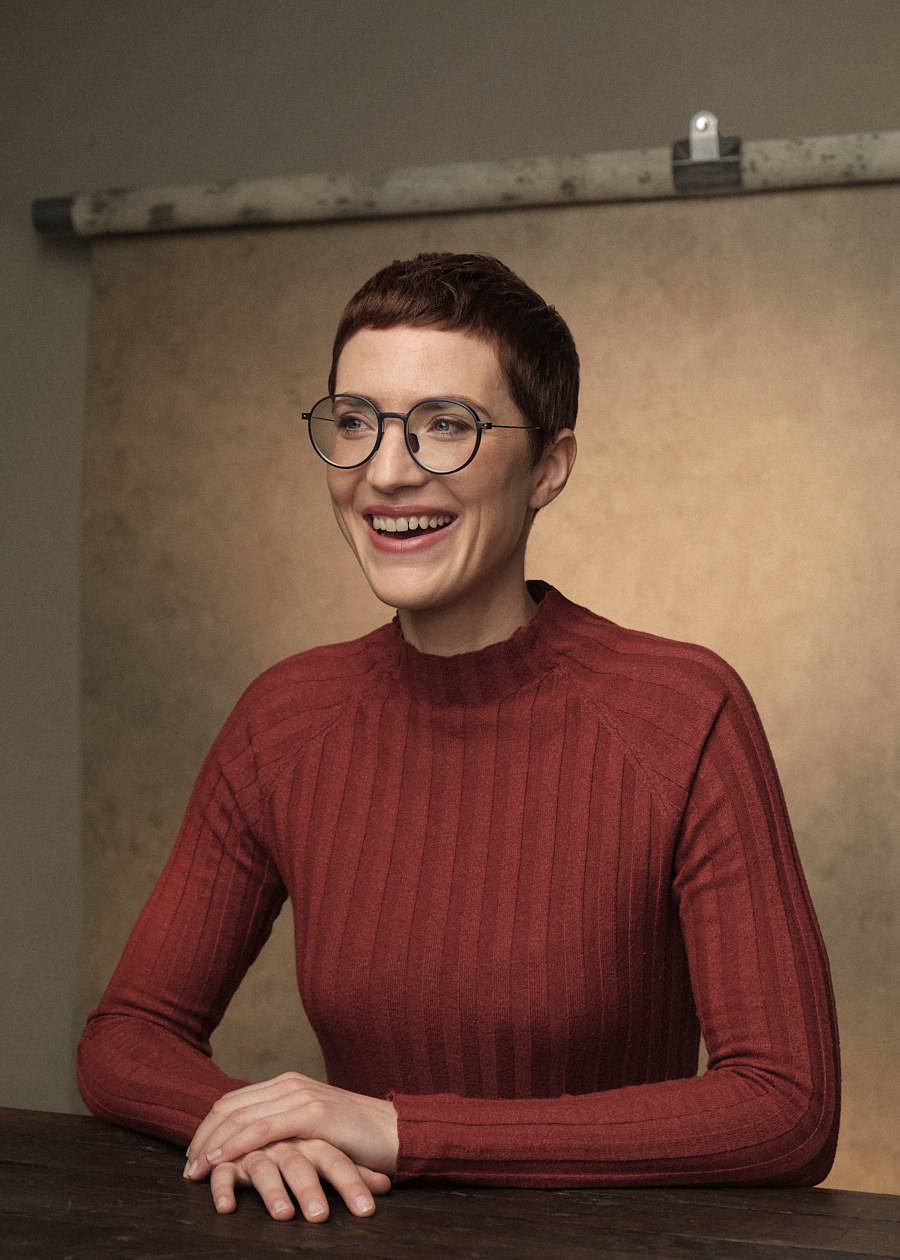 Portrait photograph of a young professional woman with short red hair and glasses, seated at a brown table. She's dressed in a red jumper, and her candid laughter, as she gazes off-camera, adds a genuine touch to this captivating portrait.