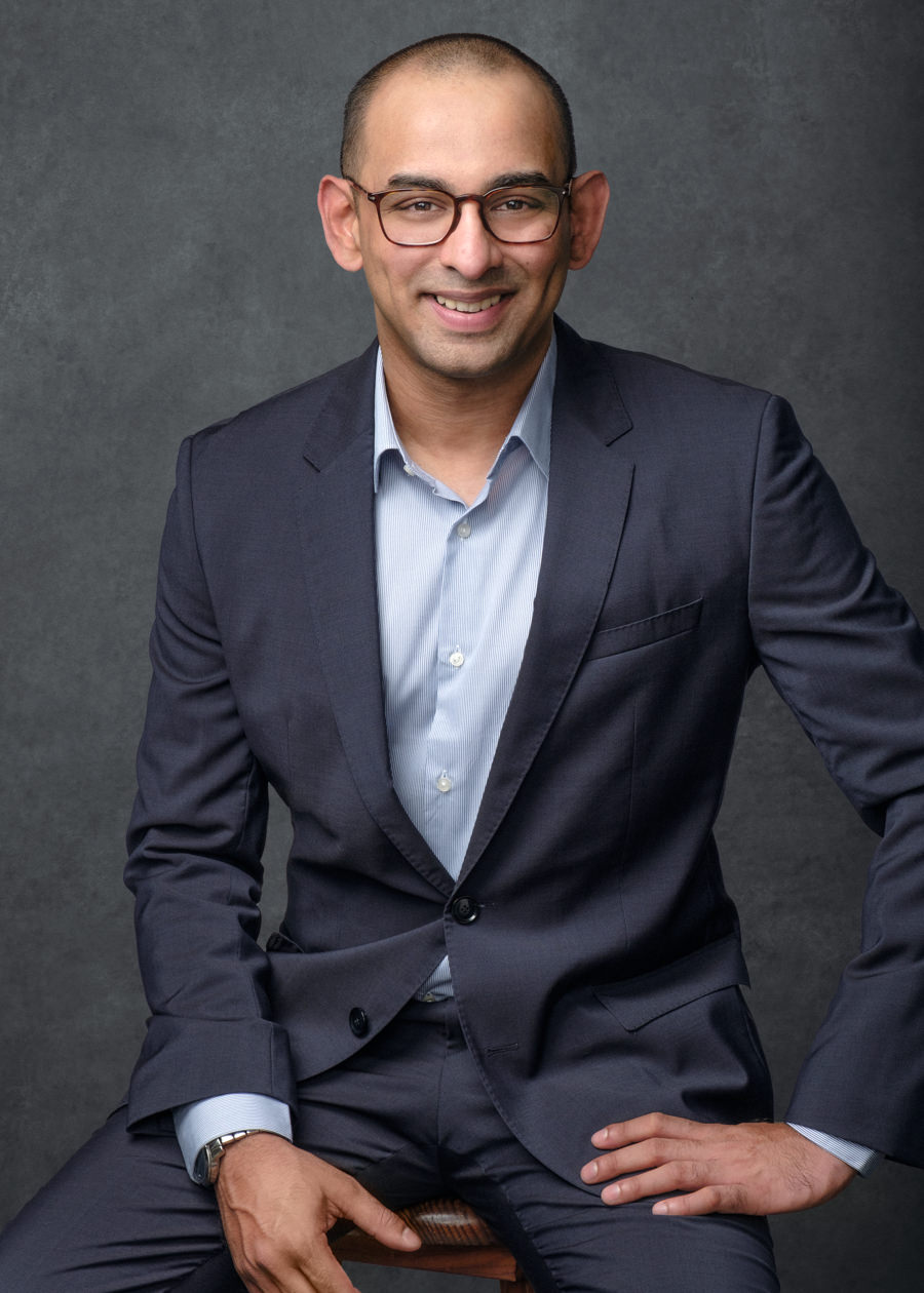 Professional portrait of an Indian businessman wearing a navy suit and blue shirt. He's sitting on a wooden stall in a very relaxed pose and is smiling naturally. He has short hair and glasses. The photo has a textured blue canvas background.