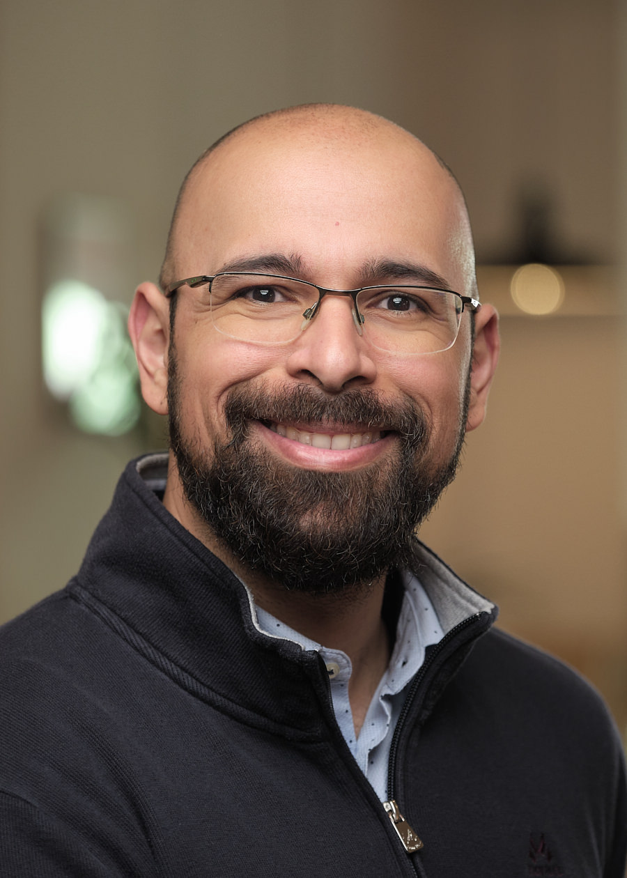 A corporate headshot of a team member from the company Mixpanel in Barcelona. The photo shots a bald man with a beard. He's wearing glasses and a blue zip up jumper with a blue shirt underneath. He's smiling. The background shows an out of focus office.