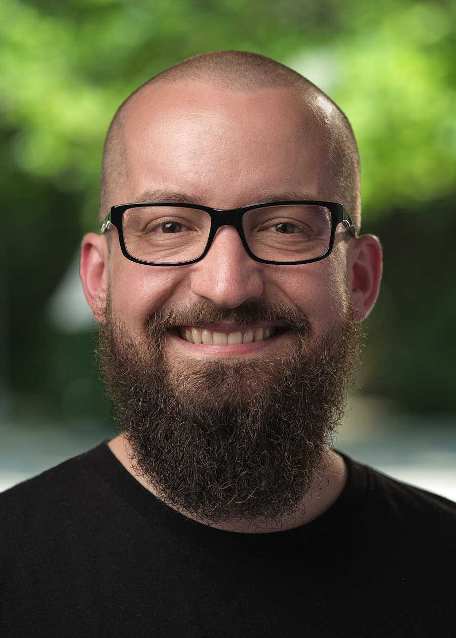A corporate headshot of a friendly man smiling directly at the camera. He has a shaved head and black glasses. He has a long beard. The background of the photo is out of focus and of trees.