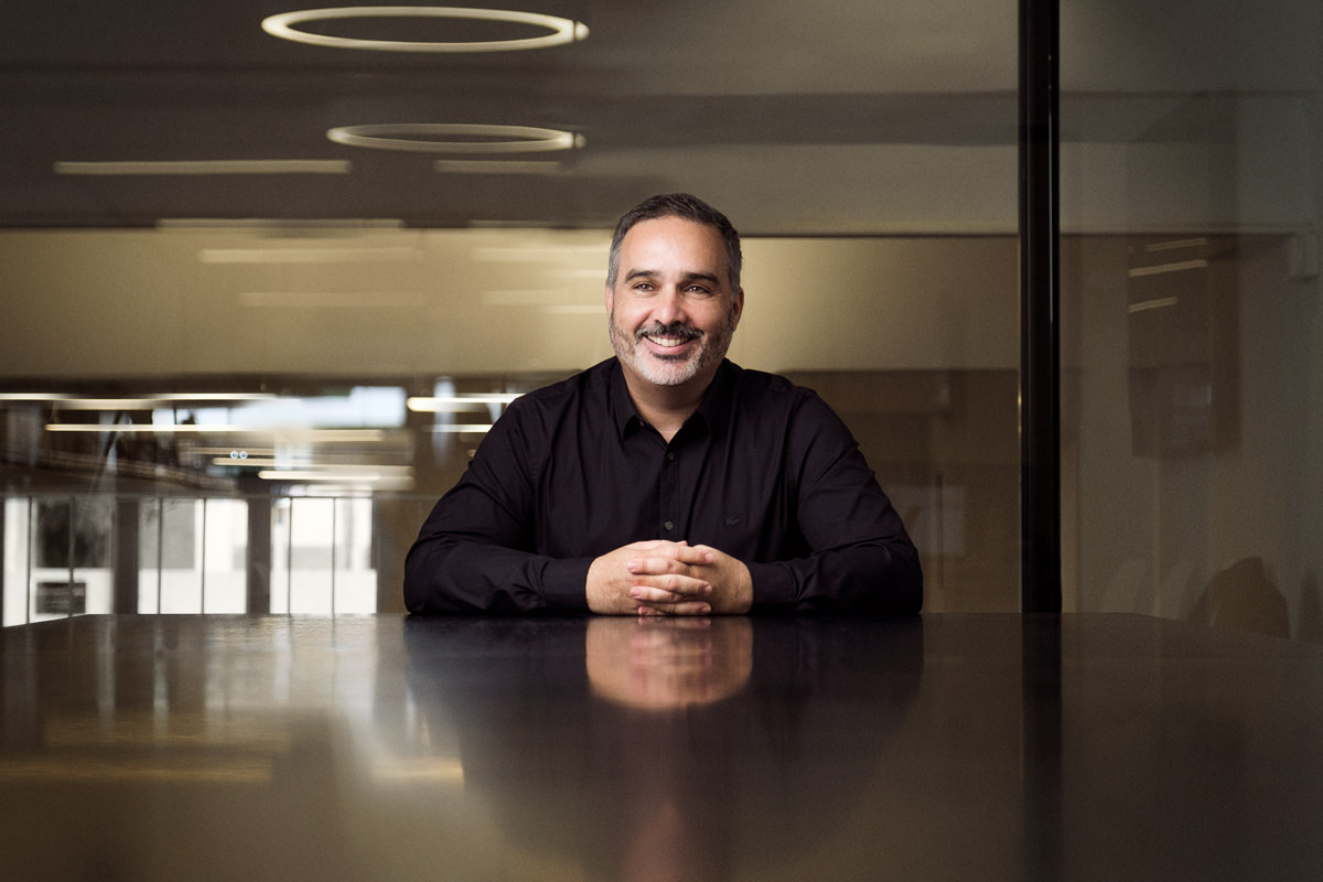 A compelling corporate portrait featuring Thomas Imfeld, the CEO of Iberent Technology. Dressed in a sleek black shirt, he sits at a meeting table in their Barcelona office, hands clasped together. His relaxed demeanor shines through as he shares a natural, hearty laugh.