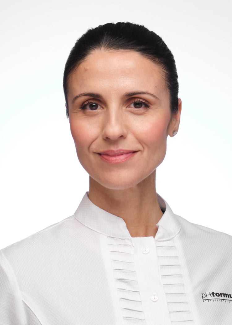 A studio portrait of a young woman who works in skincare. She's smiling confidently at the camera, whilst standing in front of a white background.